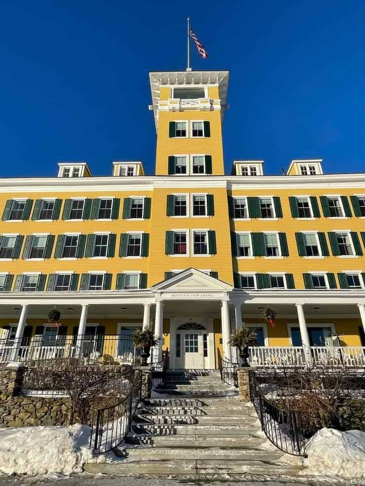 The stately facade of a yellow New England hotel under a brilliant blue sky, featuring a central watchtower with an American flag, provides a quintessential setting for winter getaways, complete with snow-dusted steps and seasonal decorations.