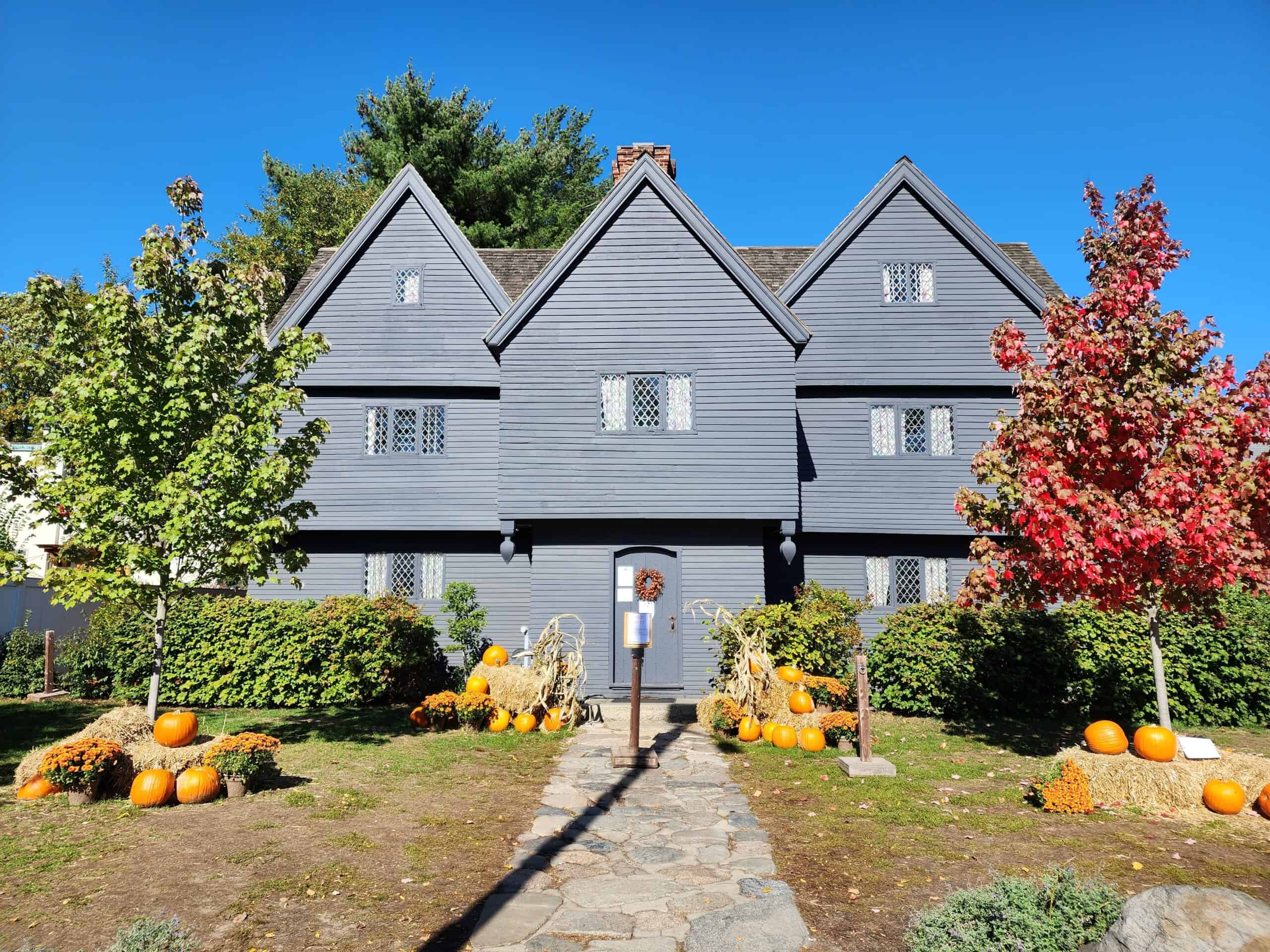 A historic dark wood-paneled house in New England, decorated with autumnal pumpkins and hay bales, captures the seasonal charm on a driving tour. The bright fall foliage provides a colorful contrast to the classic architecture, inviting visitors to explore the region's heritage