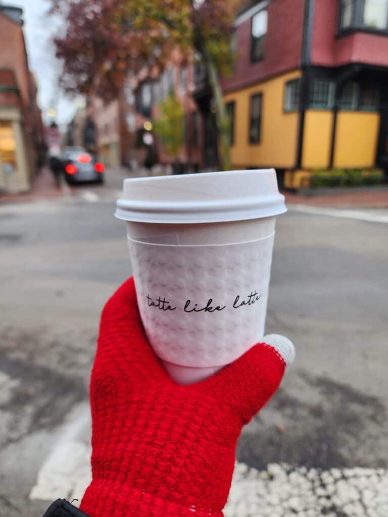 A hand in a red mitten holding a 'Tatte Bakery & Cafe' cup of coffee on a charming Boston street, capturing the essence of a cozy breakfast outing