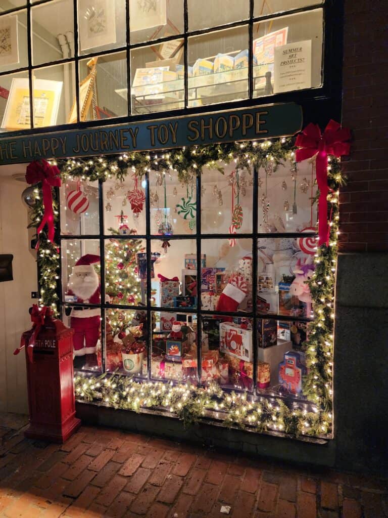 The enchanting window display of 'The Happy Journey Toy Shoppe' adorned with festive Christmas lights and red bows, capturing the holiday spirit in New England Christmas towns. The window beckons with a variety of classic toys and holiday decorations, creating a magical streetscape scene straight out of a storybook