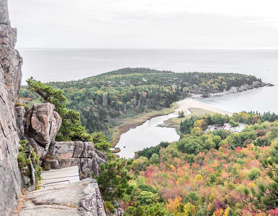 A breathtaking aerial view from a cliffside, overlooking a tapestry of fall foliage in New England, with a backdrop of a calm bay meeting the Atlantic Ocean. This panoramic vista captures the essence of exploring New England, where land and sea meet amidst the splendor of autumn.