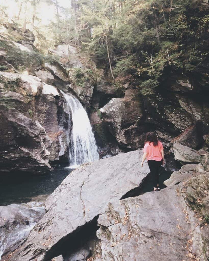 A person in a pink shirt climbs over rocky terrain toward a serene waterfall nestled in a forest, a moment of adventure and natural beauty to be found in the wilderness
