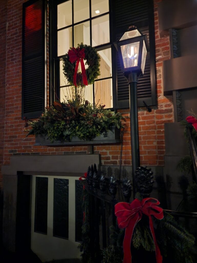 A warm, glowing gas lantern casts a soft light on a festive wreath with a bright red bow, adorning a window of a brick building with elegant greenery and ribbons, evoking a quaint and cozy holiday scene.