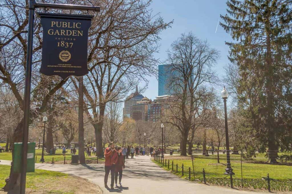 Visitors strolling through the Boston Public Garden under a clear blue sky, with the park's signature blue and gold sign in the foreground, a peaceful activity among the many things to do in Boston