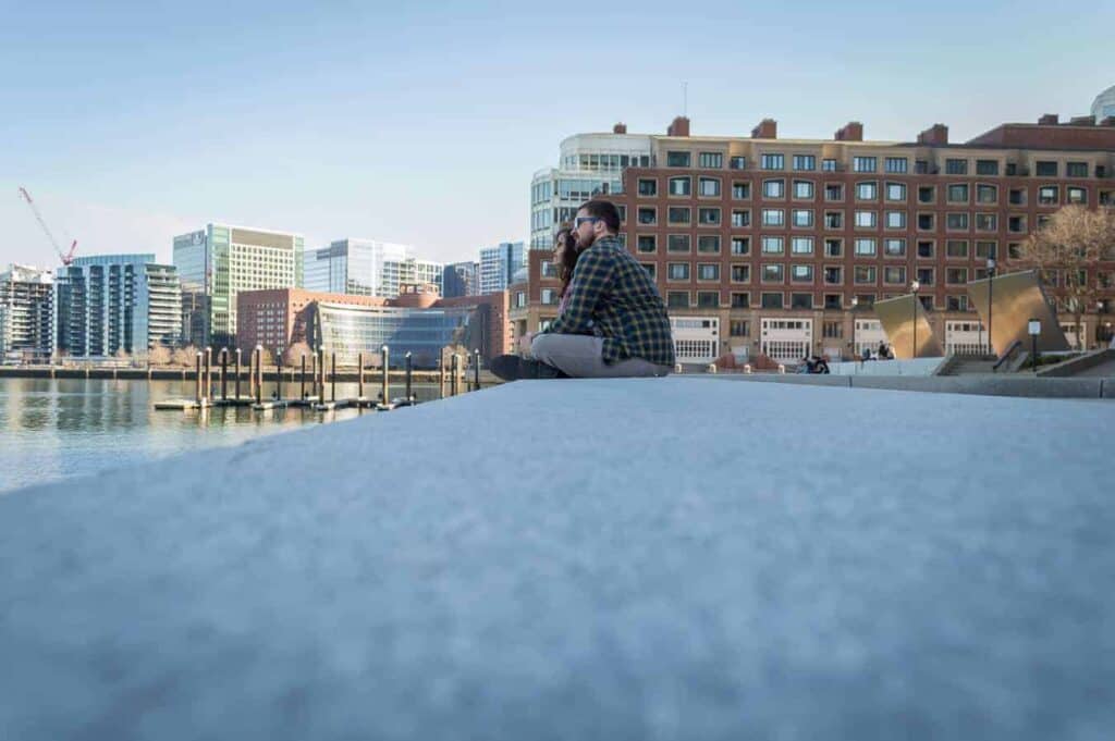 A thoughtful moment captured on a serene Boston waterfront, with a man and woman sitting on the edge contemplating the view, a peaceful scene to enjoy on a Boston trip