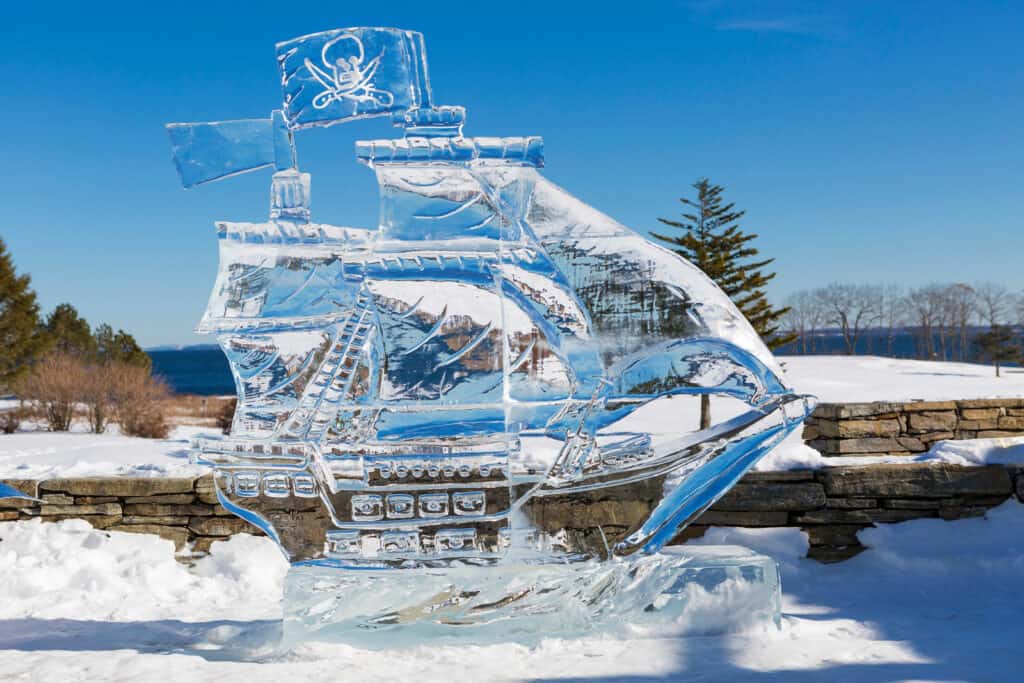 GPT
An intricate ice sculpture of a pirate ship is showcased outdoors, with its detailed masts and sails glistening under a clear blue sky. The sculpture stands on a stone wall against a backdrop of snowy terrain and leafless trees, with a calm lake visible in the distance
