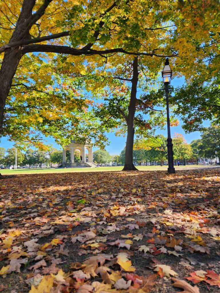 A gorgeous sunny fall day in salem ma in october. Fall leaves lay all over the ground while a white gazebo in a park is seen I'm the background and a classic lamppost nearby