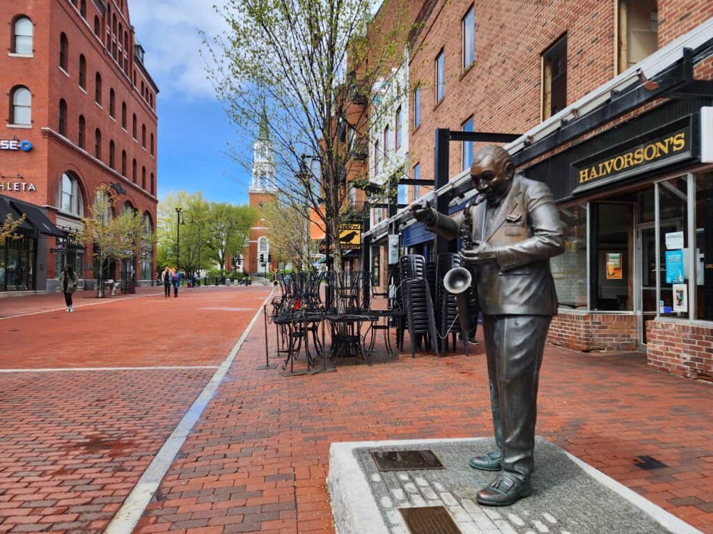 A life-size bronze statue of a saxophonist on the vibrant, brick-lined Church Street Marketplace in Burlington, VT, captures the essence of the city in the fall with hints of autumnal foliage in the backdrop.