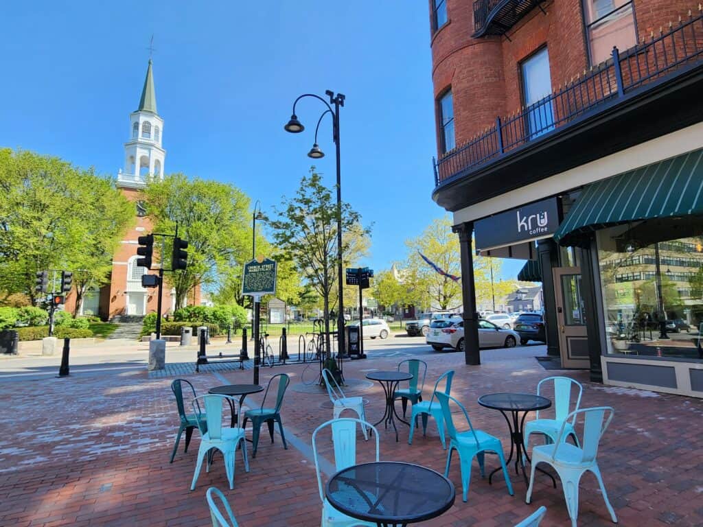 Outdoor seating at a café in Burlington, Vermont, featuring turquoise chairs on brick pavement, with the iconic steeple of the First Unitarian Universalist Church rising in the background under a clear blue sky
