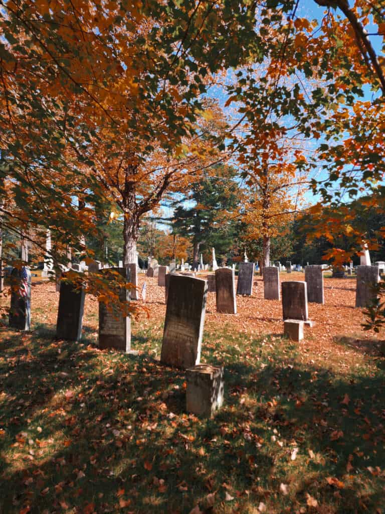An old New England cemetery in autumn, where historical gravestones stand among a carpet of fallen leaves, with trees dressed in the quintessential reds and oranges of the season.