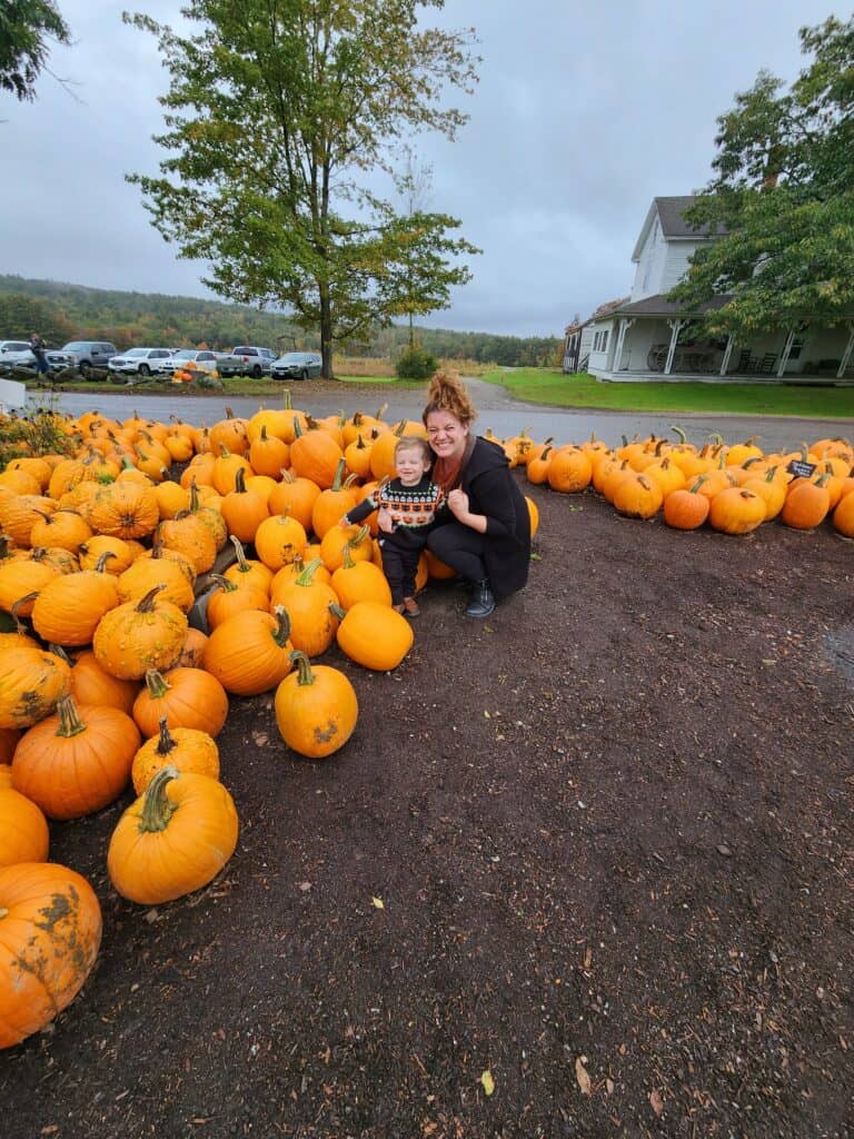 A smiling woman and child crouch among a large spread of bright orange pumpkins at a New England pumpkin patch, with lush green trees and a cloudy sky in the background, encapsulating the festive spirit of the region's fall season.