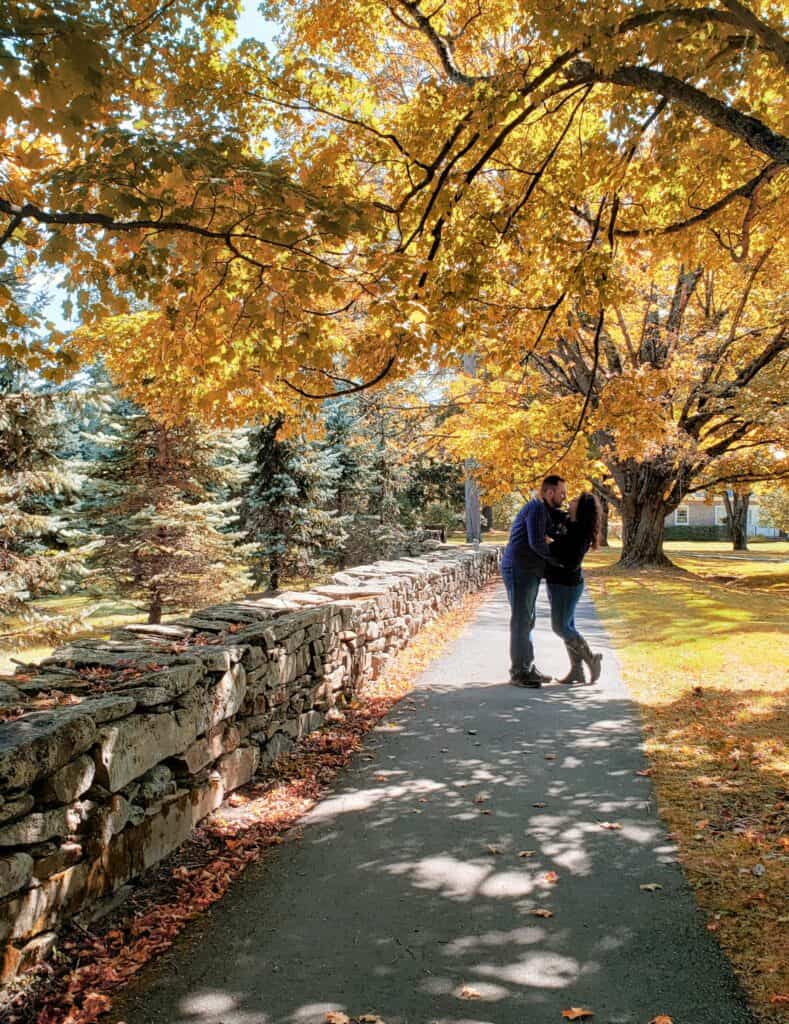 A man and a woman embrace among fall foliage next to a stone wall in Vermont
