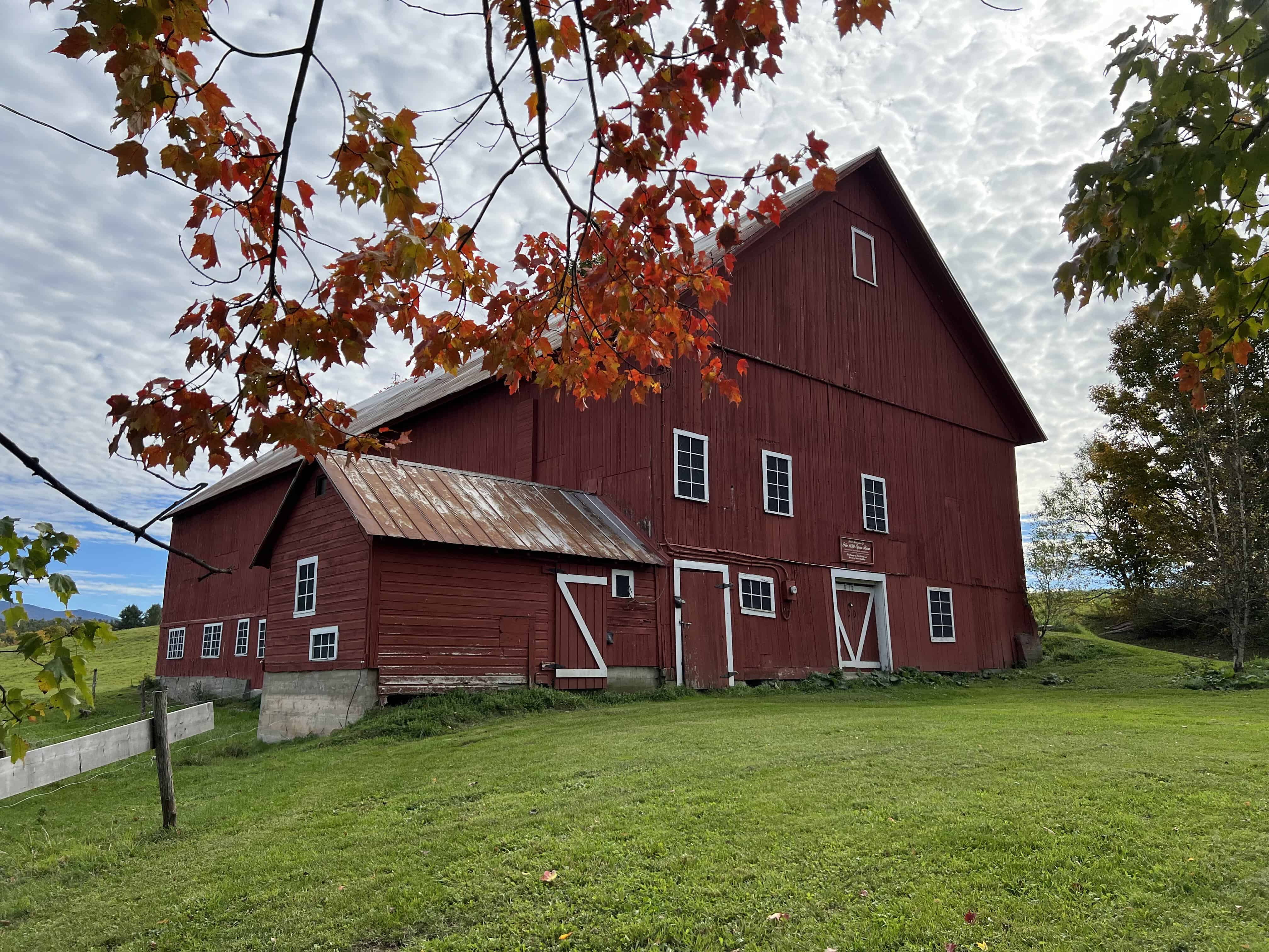 A traditional red barn in New England surrounded by a verdant lawn with autumnal foliage. The changing leaves add a pop of orange and red against the backdrop of a cloudy sky